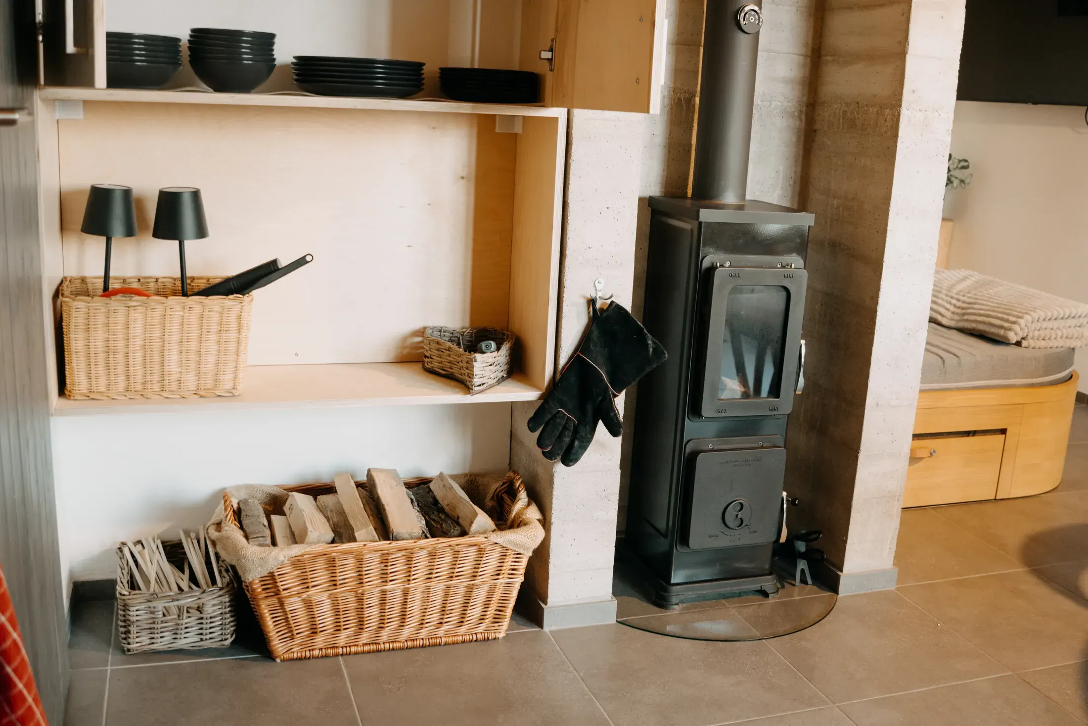 The wood-burning stove beside shelves of crockery and baskets of split logs.